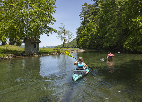 Two people in kayaks paddling back up the Susquehanna River to Otsego Lake on a beautiful sunny day.